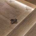 Im Untergeschoss verschmelzen beige- und braunfarbener Travertin, edles Nussholz, metallische Strukturen und feines Leder zu einer harmonischen Einheit. Das Zusammenspiel aus klaren Linien und sanft gerundeten Formen prägt dabei maßgeblich die Atmosphäre und das Geschehen. Angrenzend zur Tanzfläche wurden insgesamt vier massive Sekttische, bestehend aus Travertin Romano Classico (Unterteil) und Travertin Titanium (Oberteil) positioniert, die zum entspannten Verweilen einladen. In Präzisionsarbeit wurden in die Sekttische Vertiefungen sowie konische Aussparungen für Sektkübel eingearbeitet.