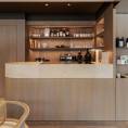 ####Die Bar aus Travertin Romano Classico bildet einen ruhigen Kontrast zu den Holzflächen und betont die reduzierte Formsprache des Raums. Das Element bietet einen Ort, an dem Besucher während des Einkaufserlebnisses in entspannter Atmosphäre einen Kaffee genießen können.