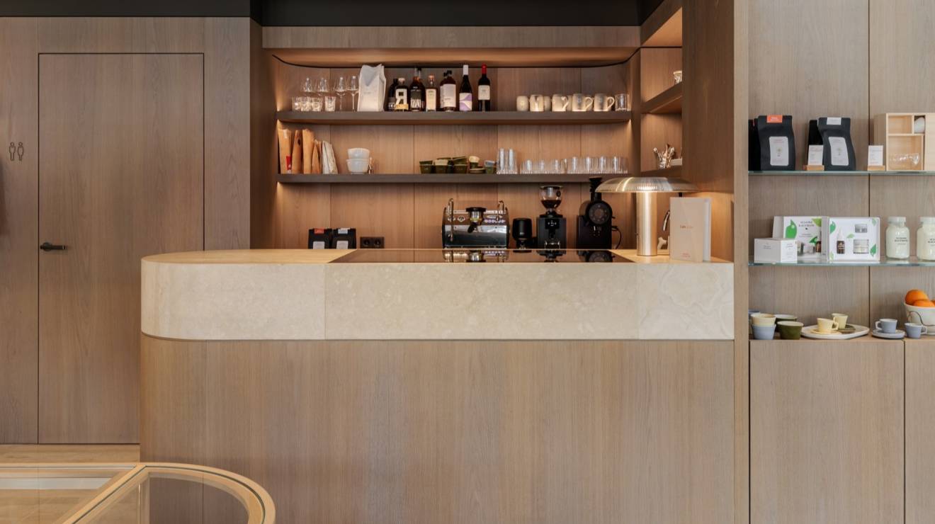 ####Die Bar aus Travertin Romano Classico bildet einen ruhigen Kontrast zu den Holzflächen und betont die reduzierte Formsprache des Raums. Das Element bietet einen Ort, an dem Besucher während des Einkaufserlebnisses in entspannter Atmosphäre einen Kaffee genießen können.