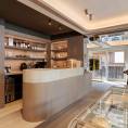 ####Die Bar aus Travertin Romano Classico bildet einen ruhigen Kontrast zu den Holzflächen und betont die reduzierte Formsprache des Raums. Das Element bietet einen Ort, an dem Besucher während des Einkaufserlebnisses in entspannter Atmosphäre einen Kaffee genießen können.