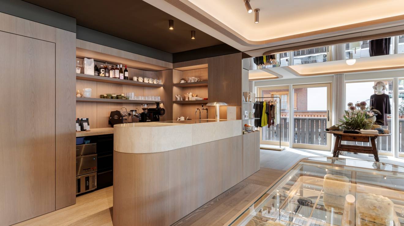 ####Die Bar aus Travertin Romano Classico bildet einen ruhigen Kontrast zu den Holzflächen und betont die reduzierte Formsprache des Raums. Das Element bietet einen Ort, an dem Besucher während des Einkaufserlebnisses in entspannter Atmosphäre einen Kaffee genießen können.
