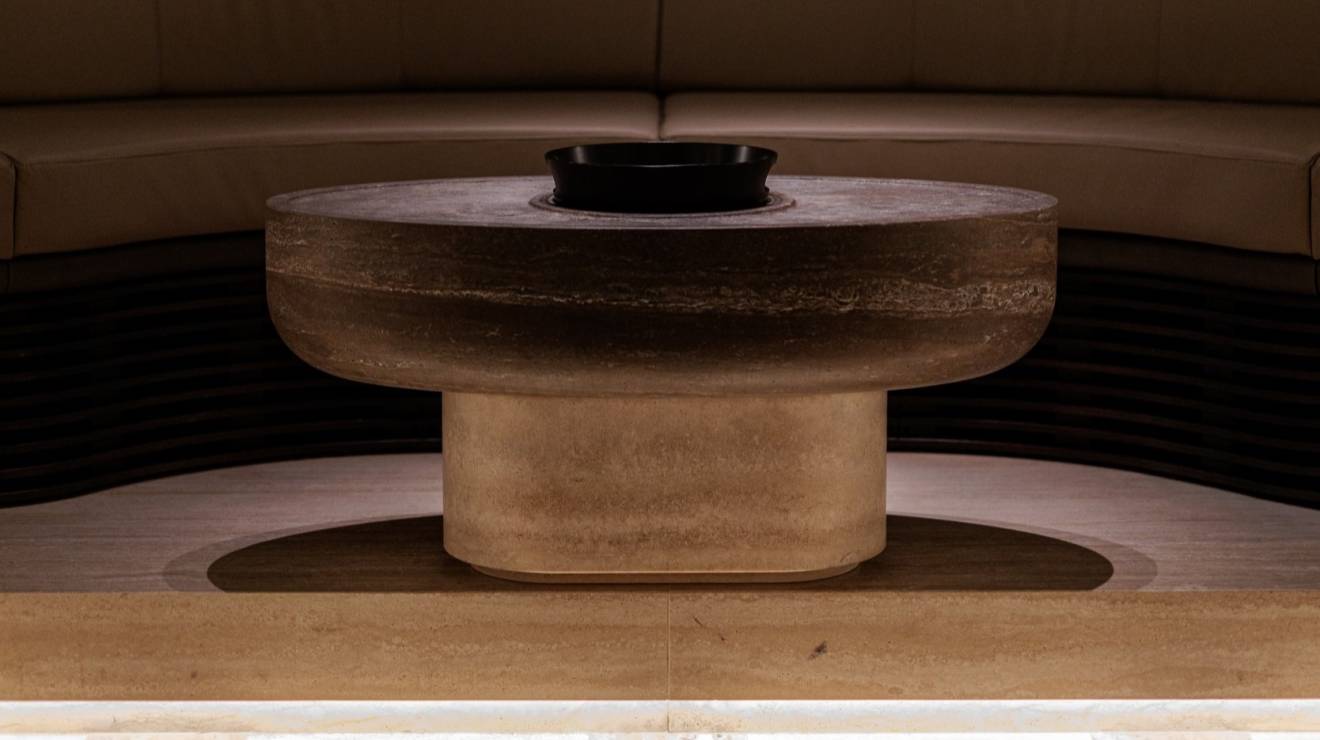 ####Im Untergeschoss verschmelzen beige- und braunfarbener Travertin, edles Nussholz, metallische Strukturen und feines Leder zu einer harmonischen Einheit. Das Zusammenspiel aus klaren Linien und sanft gerundeten Formen prägt dabei maßgeblich die Atmosphäre und das Geschehen. Angrenzend zur Tanzfläche wurden insgesamt vier massive Sekttische, bestehend aus Travertin Romano Classico (Unterteil) und Travertin Titanium (Oberteil) positioniert, die zum entspannten Verweilen einladen. In Präzisionsarbeit wurden in die Sekttische Vertiefungen sowie konische Aussparungen für Sektkübel eingearbeitet.