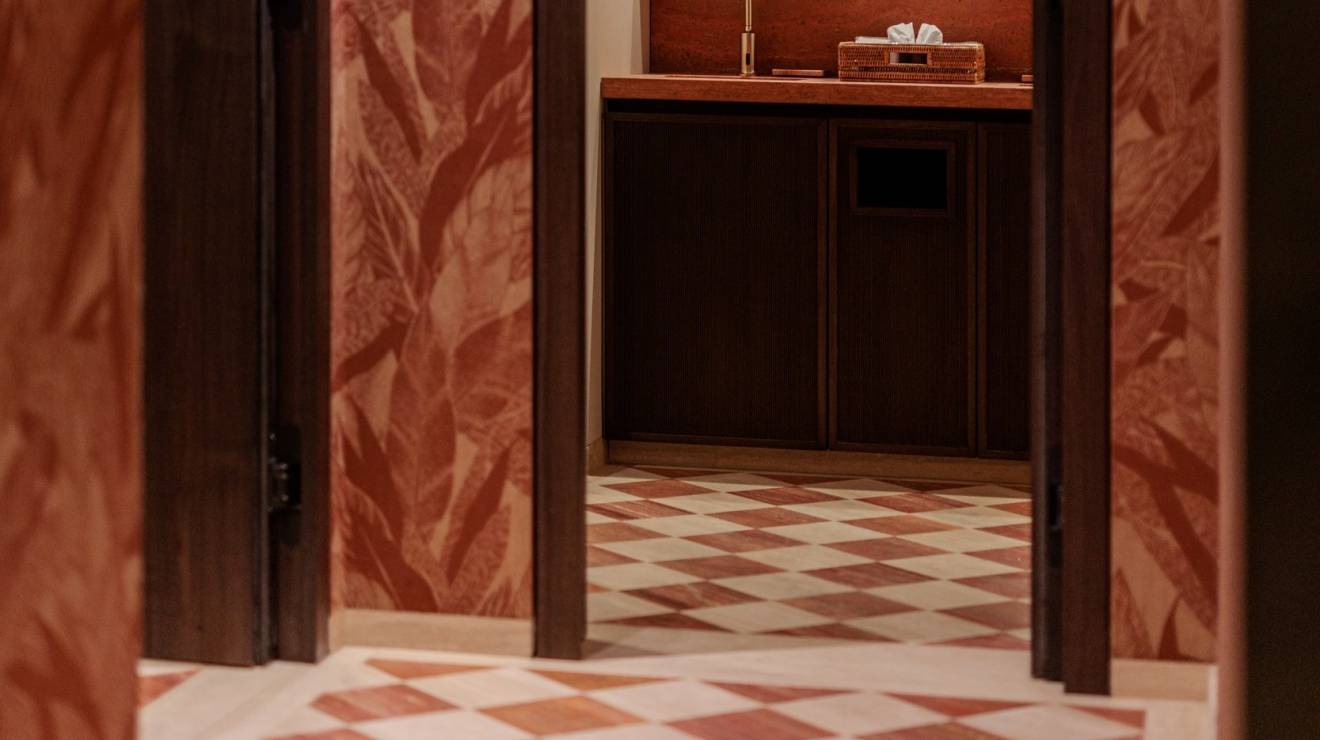 ####Der WC-Bereich bringt Farbe in die Räumlichkeiten. Die rotfarbenen Akzente setzen einen markanten, aber harmonischen Kontrast zur beige-braunen Farbgebung des Barbereichs. Bestehend aus knapp 60 verschiedenen Teilen, erzeugt die aufwendige Diagonalverlegung des Bodenbelags durch die Farbkombination von rotem und beigen Travertin ein mediterranes Flair. <p>
Abgerundet wird das Erscheinungsbild des Bereichs durch die ebenfalls in rotem Travertin ausgeführten Wandplatten und Waschtische. Letztere wurden, um dem massiven Stil der Architektur gerecht zu werden, aus einzelnen, in Gehrung verleimten Platten gefertigt. <p>